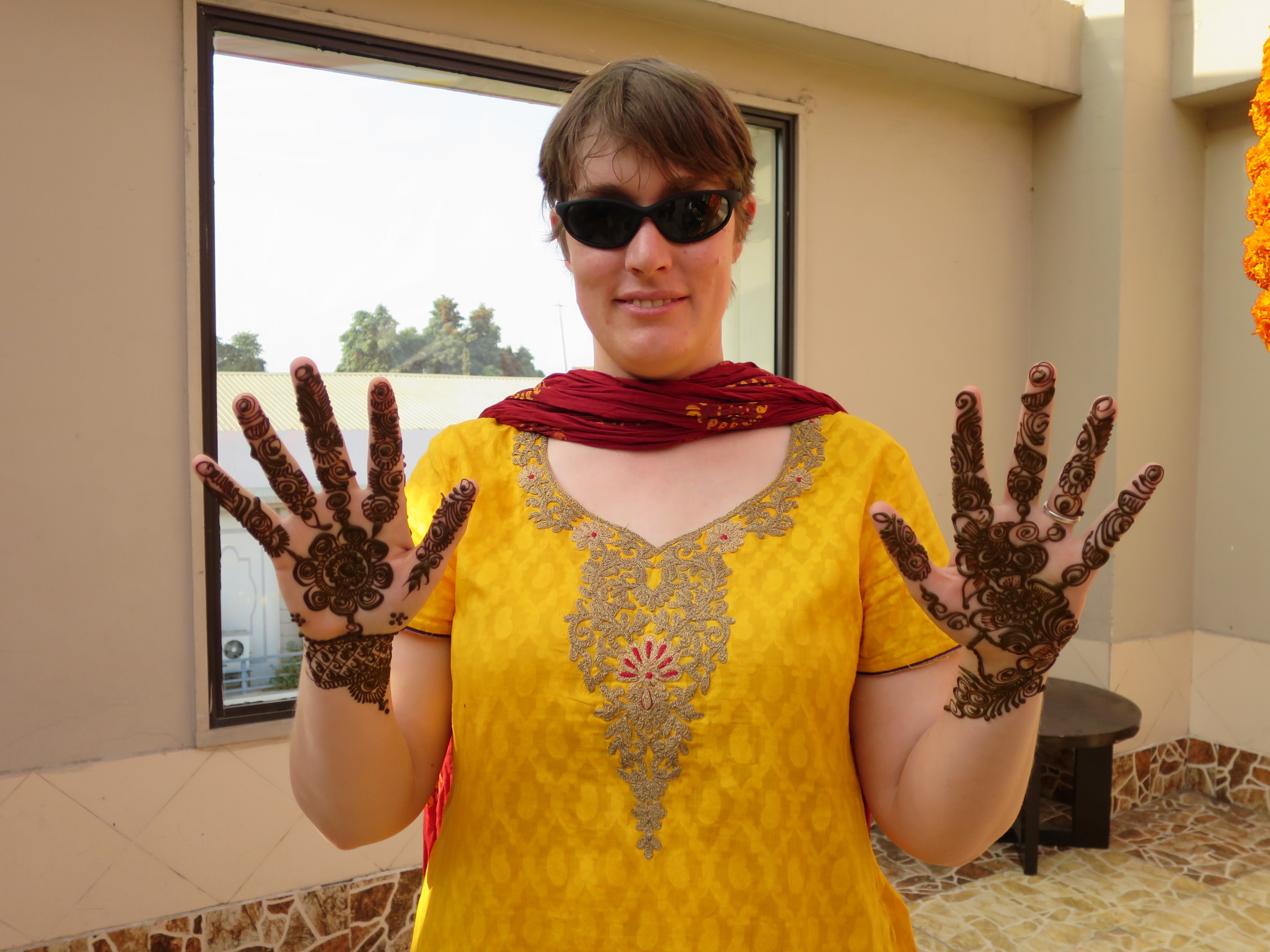 Katie, a white woman, is in the center pictured from mid-chest to the top of her forehead. She has short brown hair and is wearing black sunglasses and a slight smile. She is wearing a red scarf with dark yellow pattern backwards on her neck as a dupata. Her Indian top is bright yellow adorned with gold and dark red embroidery. She is showing the palms of her hands which have an elaborate henna design on each from four inches below the wrist to the tips of her fingers. The designs on both hands are different. She is wearing a silver wedding ring.