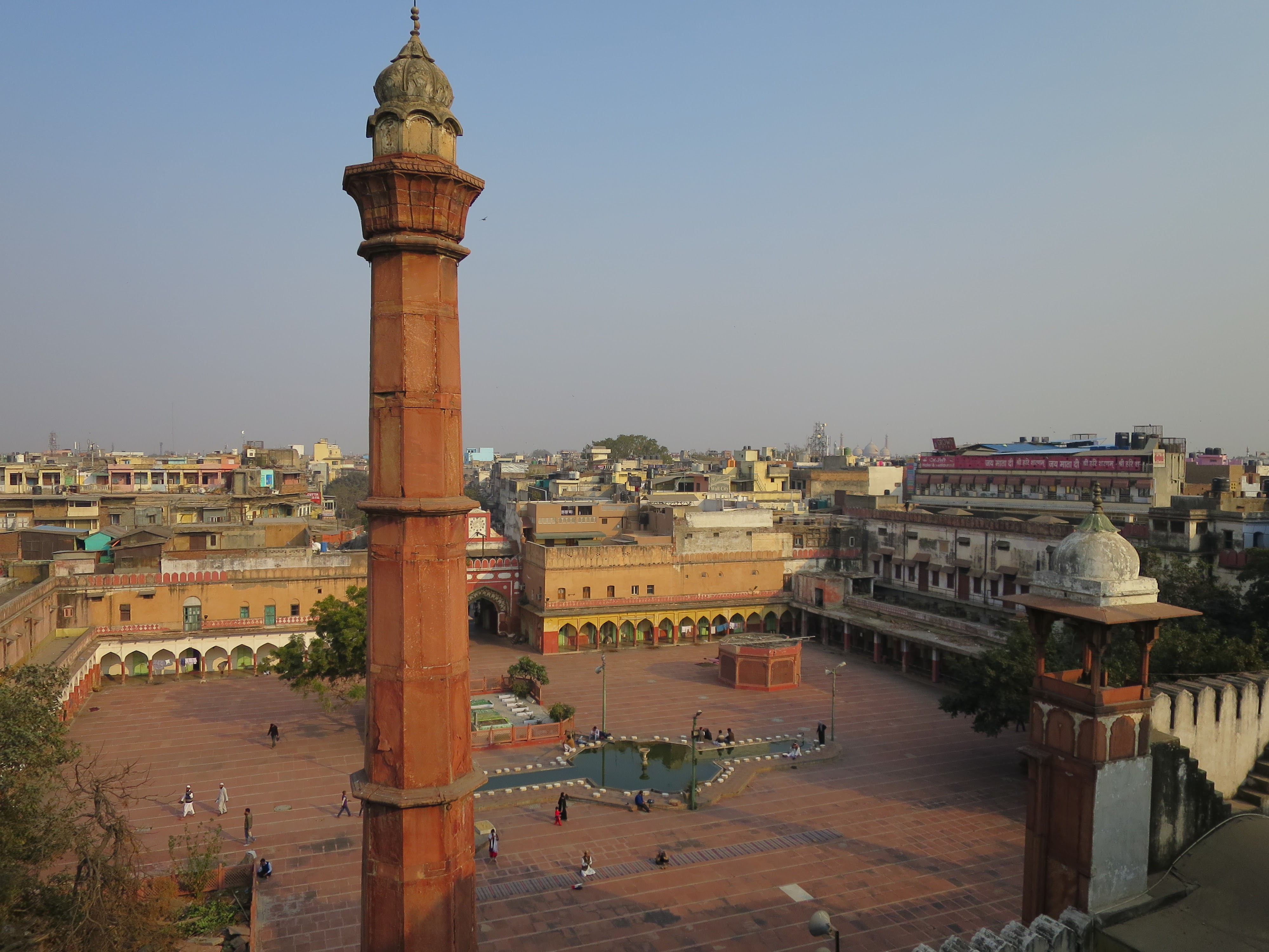 The photographer is atop a 4 story building, looking down on the city of Delhi. There is a large clay red tower in the foreground. Beyond that is a clay red square with several trees. The two story buildings surrounding the square have dozens of one story arches leading onto the square. The colors in the picture are mainly tans, browns, tellows, clay red, and all bright colors are muted by a layer of dust. The sky has no clouds but is more gray than blue.