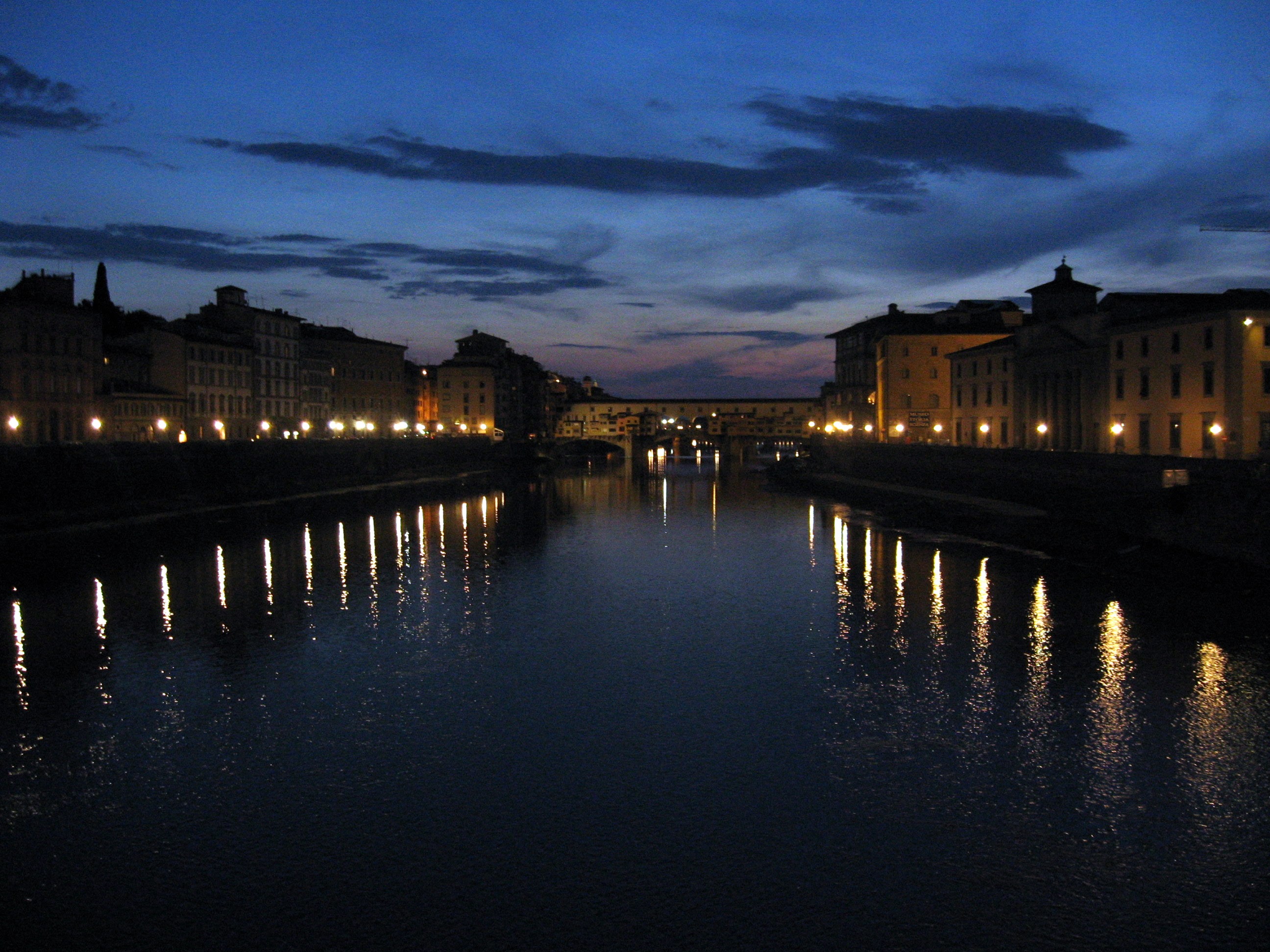 The photographer is standing on a bridge at night, looking down the Arno at the Ponte Vecchio. The streets on both sides of the river have street lights that are reflected in the dark water. The Ponte Vecchio is a bridge that has shops on it, so there are windows and several lights where it crosses the river. The sky is dusky blue with some clouds.