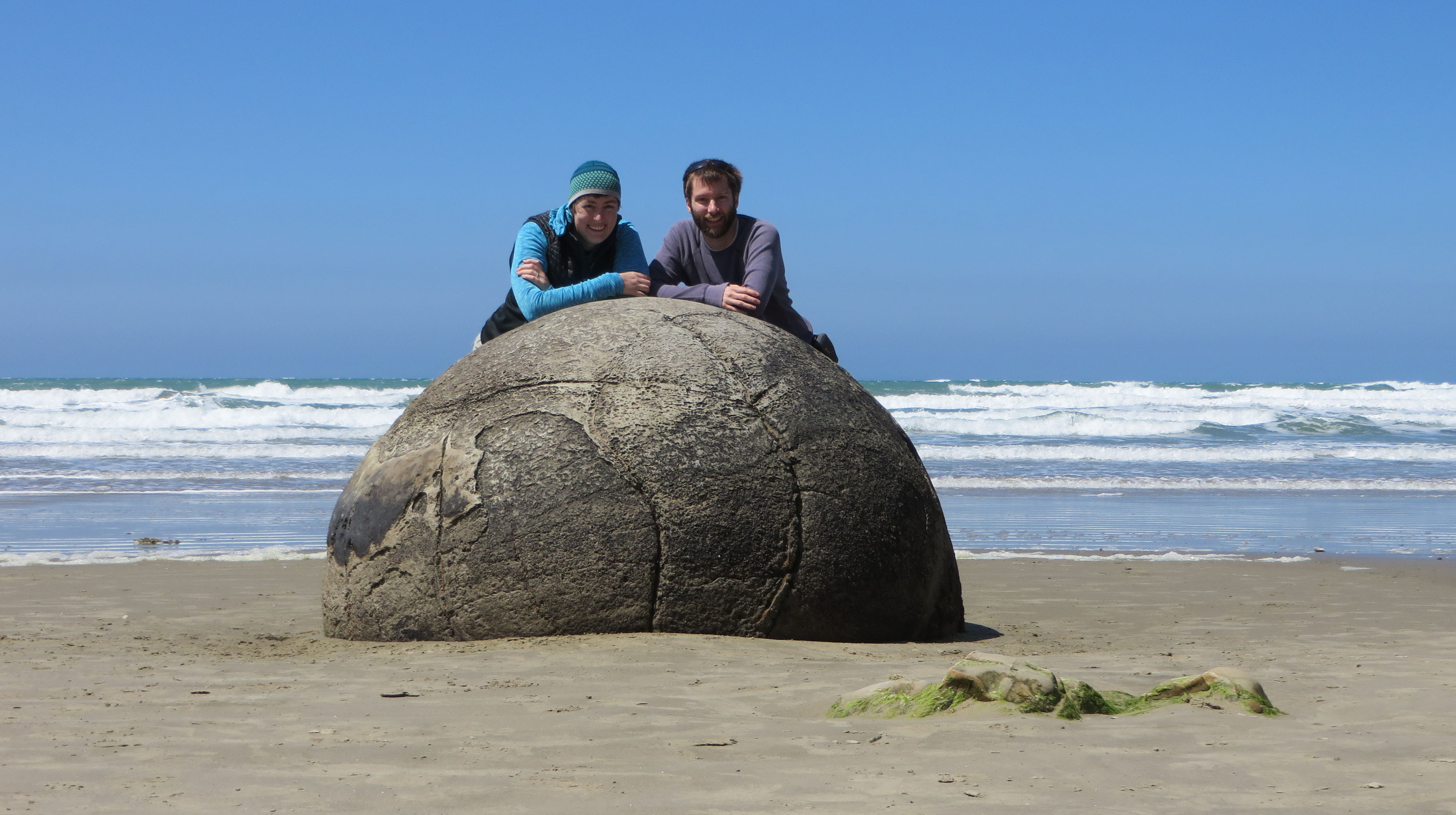 Katie, a white woman on the left, and Ben, a white man on the right, are leanding on a giant spherical boulder close together. Katie is wearing a light blue long sleeved top, a black vest, and a teal and seafoam green pattered hat. Ben is wearing a grayish-purple long sleeve top with sunglasses on the top of his head. He has brown hair and a short, trim beard and mustache. Both people are smiling. Most of the foreground is sand, but on the rightside is an oddly shaped rock with some lime green moss. In the background is the ocean and you can see several sets of docile waves rolling in.