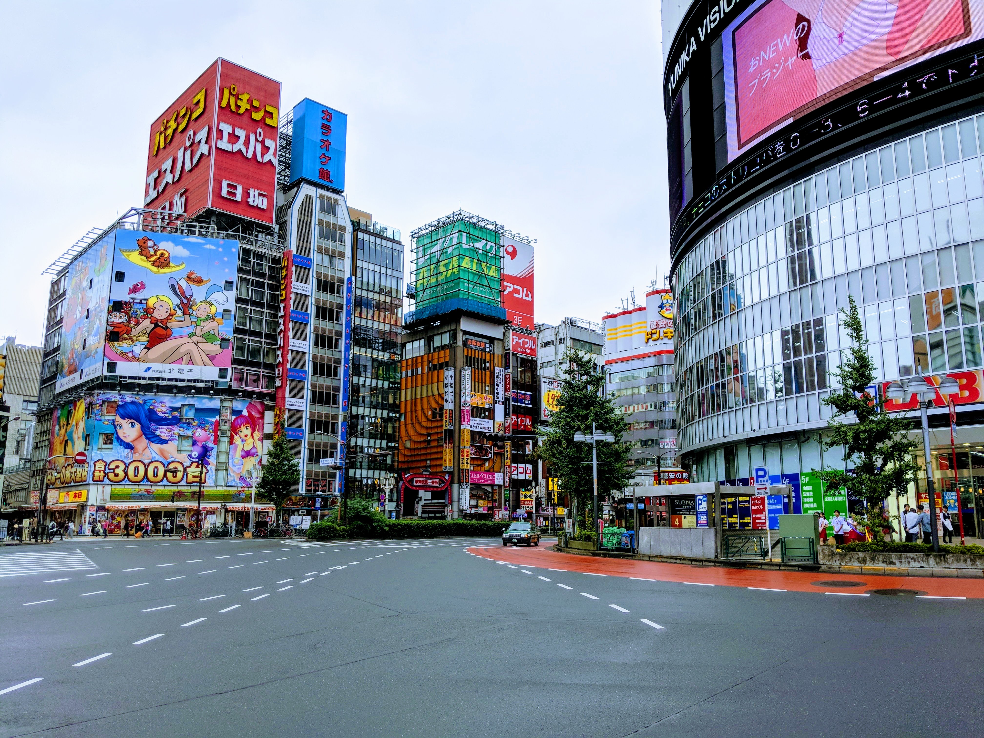 A busy intersection in Tokyo is written. There are two billboards and a giant sign on the left. The sign is atop a 9 story building and is probably 6 stories tall. It has three lines of text in Japanese. The two billboards below it have cartoonish people and animals. More bright signs and text are on all the remaining buildings. There is a lot of blue, red, orange, and green. There is only one car on the road and fewer than 20 small people on the sidewalks.